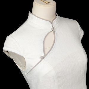May include: A white sleeveless dress featuring a mandarin collar with light grey piping trim. The bodice includes a teardrop-shaped cutout at the neckline, secured by two small, round, fabric-covered buttons in a matching light grey tone.