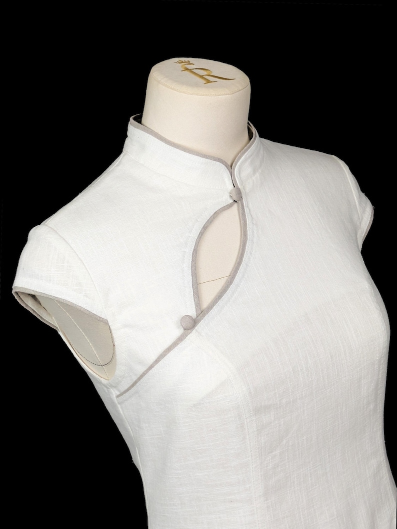 May include: A white sleeveless dress featuring a mandarin collar with light grey piping trim. The bodice includes a teardrop-shaped cutout at the neckline, secured by two small, round, fabric-covered buttons in a matching light grey tone.