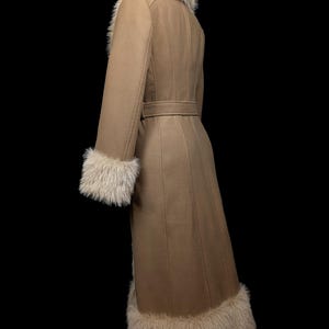 May include: A tan-colored coat with faux fur trim on the collar, cuffs, and hem. The coat features a belted waist and a long, flowing design. The coat is made of a soft, warm material, perfect for cold weather.