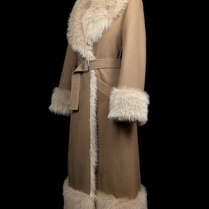 May include: A long, tan coat with faux fur trim on the collar, cuffs, and hem. The coat features a matching belt with a silver buckle. The coat is displayed on a mannequin against a black background.