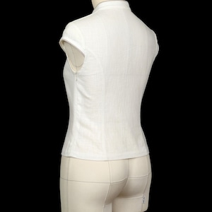 May include: A white sleeveless top with a high collar and a side zip fastening. The garment features a fitted silhouette with cap sleeves and a textured fabric finish, displayed on a mannequin against a solid black background.