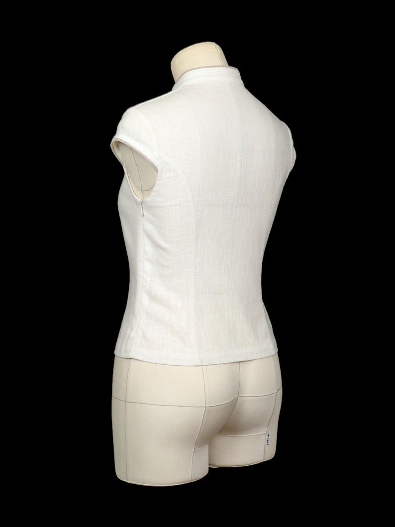 May include: A white sleeveless top with a high collar and a side zip fastening. The garment features a fitted silhouette with cap sleeves and a textured fabric finish, displayed on a mannequin against a solid black background.