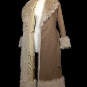 May include: A long, brown coat with faux fur trim on the collar, cuffs, and hem. The coat has a tailored fit, a belt, and a light-colored lining. The coat is displayed on a mannequin against a black background.