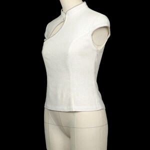 May include: A sleeveless white top featuring a mandarin collar, a keyhole cutout at the neckline, and a side zip fastening. The garment is constructed with a textured fabric and includes a small button detail near the collar and shoulder area.