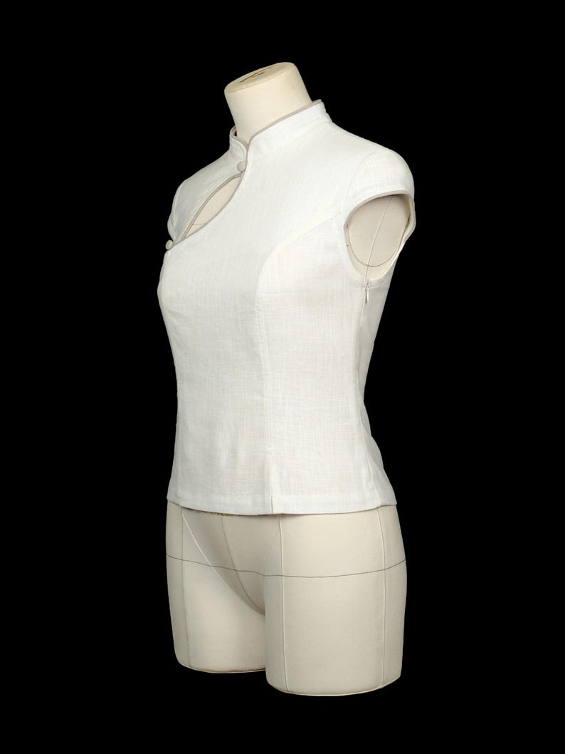 May include: A sleeveless white top featuring a mandarin collar, a keyhole cutout at the neckline, and a side zip fastening. The garment is constructed with a textured fabric and includes a small button detail near the collar and shoulder area.