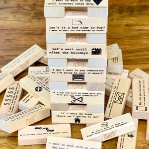 May include: A wooden Jenga game with a stack of blocks featuring humorous real estate-themed phrases. The top block reads "I want to wait to buy until interest rates drop." The blocks are stacked on a wooden surface.