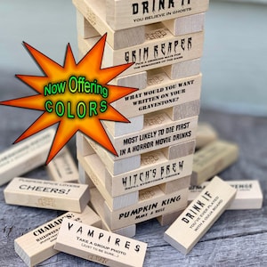 Może przedstawiać: Drewniana gra Jenga z wyzwaniami o tematyce Halloween. Klocki zawierają frazy takie jak "Wypij, jeśli wierzysz w duchy", "Grim Reaper", "Czarodziejski napój", i "Wampiry".