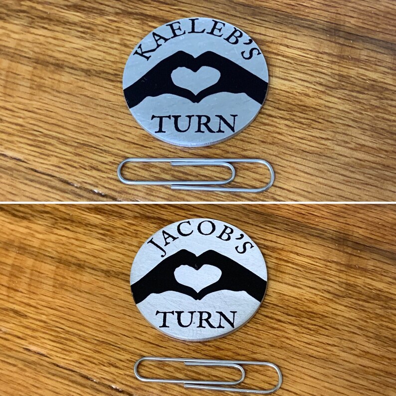 Op de afbeelding: Twee zilveren tokens met zwarte hand-silhouetten die een hartvorm vormen. De bovenste token zegt "Kaleb's Turn" en de onderste token zegt "Jacob's Turn".