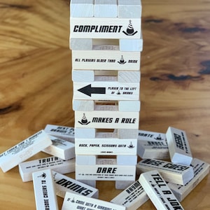 May include: A wooden block tower game with various challenges printed on each block. Some of the challenges include "Compliment", "Makes a Rule", "Dare", "Tell a Joke", and "Drinks".