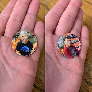 Op de afbeelding: Twee ronde, oranje en witte, gepersonaliseerde geluksbrengers met een foto van een jonge jongen. De tekst op de tokens luidt "Papa's Lucky Charm".
