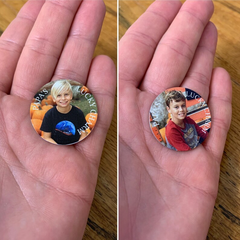 Op de afbeelding: Twee ronde, oranje en witte, gepersonaliseerde geluksbrengers met een foto van een jonge jongen. De tekst op de tokens luidt "Papa's Lucky Charm".