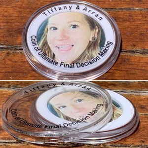 Op de afbeelding: Een doorzichtige plastic munt met een wit centrum met daarop het gezicht van een vrouw en de tekst "Tiffany & Arran Coin of Ultimate Final Decision Making".