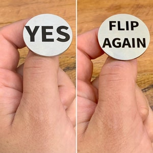 Op de afbeelding: Een zilveren munt met het woord "YES" in zwart gedrukt op de ene kant en "FLIP AGAIN" in zwart gedrukt op de andere kant.