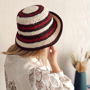 May include: A white and brown striped bucket hat with red stripes. The hat is crocheted and has a wide brim.