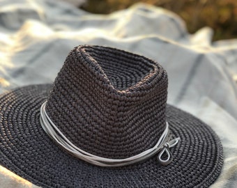Puede incluir: Un sombrero de paja tejido de color gris oscuro con una cinta blanca. El sombrero está sobre una manta blanca.