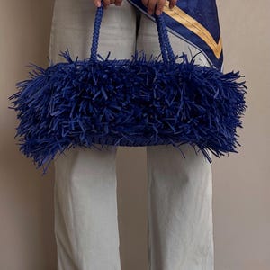 May include: A vibrant blue handbag with a fringed design. The bag features braided handles and a rectangular shape. The fringed material gives the bag a textured appearance. The bag is held by a person wearing light-colored pants.