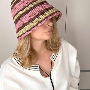 May include: A striped bucket hat in pink, brown, and yellow. The hat is made of a woven material and has a wide brim. The person is wearing a white blouse with black trim.