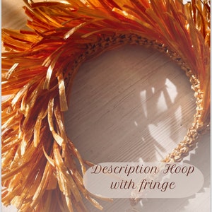 May include: A gold hoop with a fringe of thin, woven strips. The hoop is lying on a wooden surface with two gold hoop earrings next to it. The text "Description Hoop with fringe" is written on a white background.