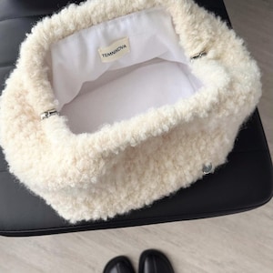 May include: Cream-colored, fluffy handbag with a soft, textured exterior. The interior is white, and the bag has silver-toned hardware. A small label inside reads "TEMNIKOVA". The bag sits on a black surface, with black boots visible below.