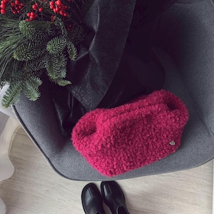 May include: A fuchsia-colored, textured clutch bag with a small silver-tone button detail. The bag is resting on a gray chair next to a bouquet of greenery and red berries. Black leather boots are at the bottom.