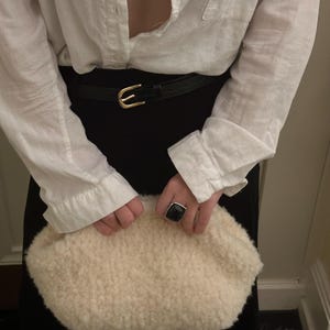 May include: Cream-colored, textured clutch held by a person. The clutch has a soft, fluffy appearance. A black belt with a gold buckle is visible. A black ring with a square, dark stone is on a finger.