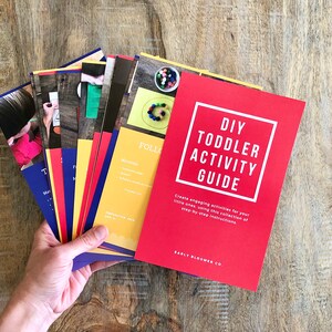 Op de afbeelding: Een rode gids met de titel "DIY Toddler Activity Guide" en de tekst "Maak leuke activiteiten voor je kleintjes met deze verzameling stapsgewijze instructies." De gids ligt bovenop een stapel kleurrijke kaarten met instructies voor verschillende activiteiten.