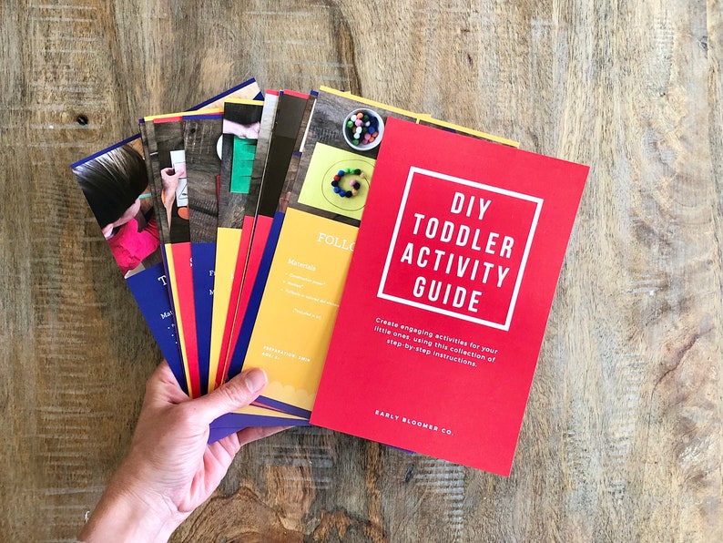 Op de afbeelding: Een rode gids met de titel "DIY Toddler Activity Guide" en de tekst "Maak leuke activiteiten voor je kleintjes met deze verzameling stapsgewijze instructies." De gids ligt bovenop een stapel kleurrijke kaarten met instructies voor verschillende activiteiten.