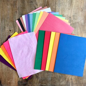 Op de afbeelding: Een assortiment gekleurde vilt- en constructiepapier vellen in verschillende tinten roze, paars, blauw, groen, geel, oranje en rood. De vellen zijn in een waaier op een houten oppervlak gerangschikt.