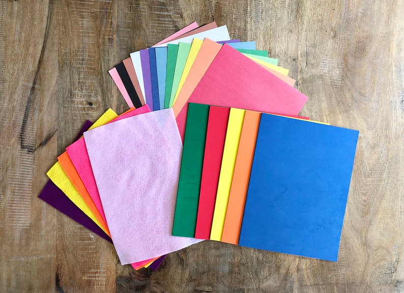 Op de afbeelding: Een assortiment gekleurde vilt- en constructiepapier vellen in verschillende tinten roze, paars, blauw, groen, geel, oranje en rood. De vellen zijn in een waaier op een houten oppervlak gerangschikt.