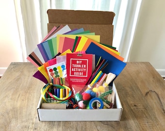 May include: A white box filled with colorful craft supplies for toddlers. The box includes construction paper, felt, pom poms, pipe cleaners, crayons, markers, and a guide for DIY toddler activities.