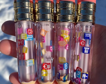 May include: Four clear lip gloss tubes shaped like pencils with pink eraser tops and gold accents. Each tube is filled with a clear gloss and colourful letter and shape charms.