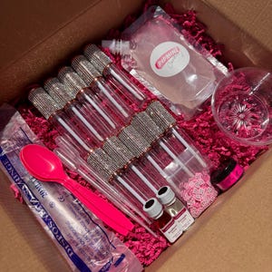 Könnte beinhalten: Ein offener Pappkarton gefüllt mit rosa Krepppapier und verschiedenen Beauty-Produkten. Enthält Lipgloss-Tuben mit Strasssteinen, einen rosa Löffel, Spritzen, kleine Flaschen und eine transparente Tasche mit dem Text "PIPMINE Cupcake".