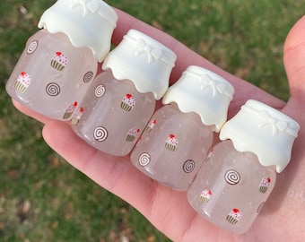 Puede incluir: Cuatro botellas pequeñas y transparentes con tapas blancas en forma de cupcake se sostienen en una mano. Las botellas están decoradas con diseños de cupcakes y espirales. Las botellas están llenas de un líquido rosa.