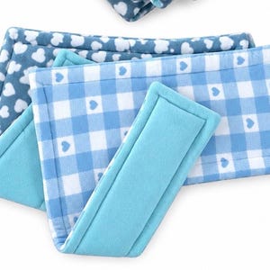 May include: A set of rectangular fabric wraps featuring a light blue and white gingham pattern with small heart icons, layered with a solid turquoise fabric wrap. The items have stitched edges and a soft, plush texture throughout the design.