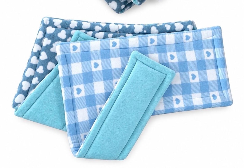 May include: A set of rectangular fabric wraps featuring a light blue and white gingham pattern with small heart icons, layered with a solid turquoise fabric wrap. The items have stitched edges and a soft, plush texture throughout the design.
