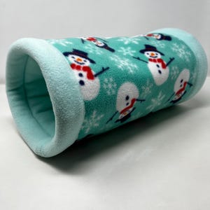 May include: A cylindrical pet tunnel made of teal fleece. The tunnel is decorated with a pattern of snowmen wearing black top hats and red scarves, along with white snowflakes. The ends of the tunnel are a lighter shade of teal.