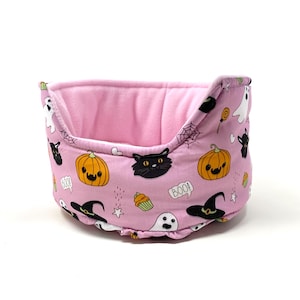 May include: A pink pet bed with a Halloween-themed pattern. The pattern includes pumpkins, black cats, ghosts, witch hats, and the word "BOO!". The interior is a solid pink color.