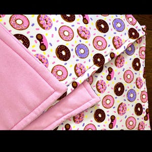 May include: A soft blanket featuring a white background with a repeating pattern of pink, brown, and purple frosted donuts with colorful sprinkles. The blanket has a solid light pink fleece backing and stitched edges throughout the design.