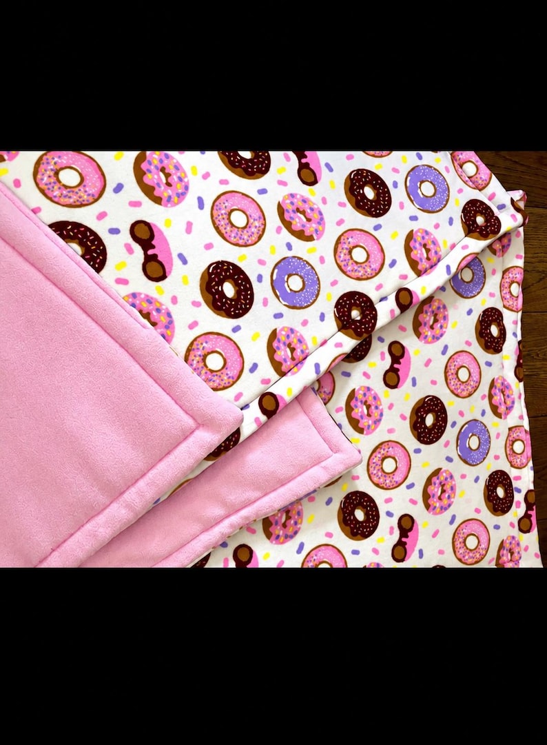 May include: A soft blanket featuring a white background with a repeating pattern of pink, brown, and purple frosted donuts with colorful sprinkles. The blanket has a solid light pink fleece backing and stitched edges throughout the design.