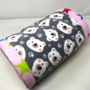 May include: A gray fleece pet bed with a repeating pattern of white polar bear faces and paw prints. The ends are pink with star and green accents. The bed is cylindrical and appears soft and comfortable.