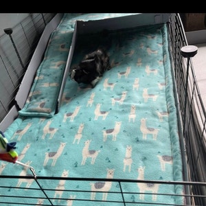 May include: A soft, light blue fleece liner for a small animal cage, featuring a repeating pattern of white and gray llamas and small green triangles. The liner includes a matching ramp cover and is enclosed within a black wire cage frame.