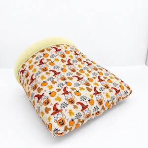 May include: A fabric pet bed with a cream-colored interior and a patterned exterior featuring cartoon cats wearing orange witch hats, holding jack-o'-lanterns, and surrounded by small orange pumpkins and scattered autumn leaves.