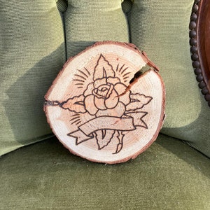 Può includere: Una fetta di legno con un disegno di una rosa bruciata a fuoco, con foglie e uno stendardo. Il legno è di colore marrone chiaro.