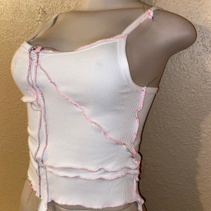 May include: White ribbed tank top with pink stitching detail. The stitching is uneven and creates a distressed look. The tank top has a scoop neck and thin straps.