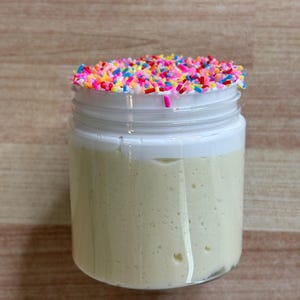 May include: A clear glass jar filled with a pale yellow substance, topped with a white layer and colourful sprinkles. The jar has a white lid and sits on a wooden surface. The contents appear to be a dessert or cosmetic product.