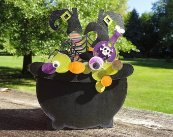 May include: A black paper cauldron decoration featuring two striped witch legs, a purple potion bottle with a skull icon, a black spider, and various colorful circular accents with googly eyes emerging from the top of the pot.