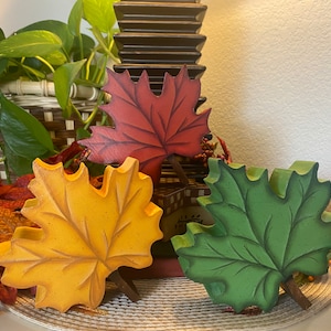 May include: Three wooden leaf decorations in fall colors: yellow, red, and green. Each leaf has a detailed, carved design and a small brown stem. The leaves are arranged with other fall-themed decor.