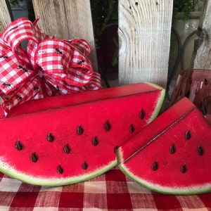 May include: Two wooden watermelon slices, one large and one small, with painted red flesh and black seeds. The slices are on a red and white checkered tablecloth. A red and white gingham bow with black ants is in the background.
