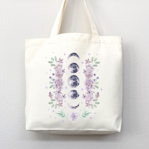 May include: A white tote bag with a design of purple flowers and a crescent moon in various phases. The design is centered on the bag.
