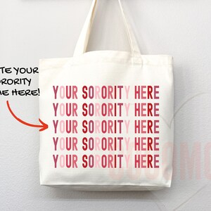 Op de afbeelding: Witte canvas draagtas met de tekst "YOUR SORORITY HERE" die vijf keer wordt herhaald in roze en rode tinten. De tekst "WRITE YOUR SORORITY NAME HERE!" staat in het zwart aan de linkerkant van de tas.